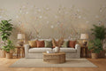 A styled interior scene showing a large wall mural with delicate floral branches, birds, and a peacock motif in soft beige, blush pink, green, and brown tones. In front of the wall is a light wood cabinet, a small chair with a draped green textile, and a table lamp on a pale wood floor.